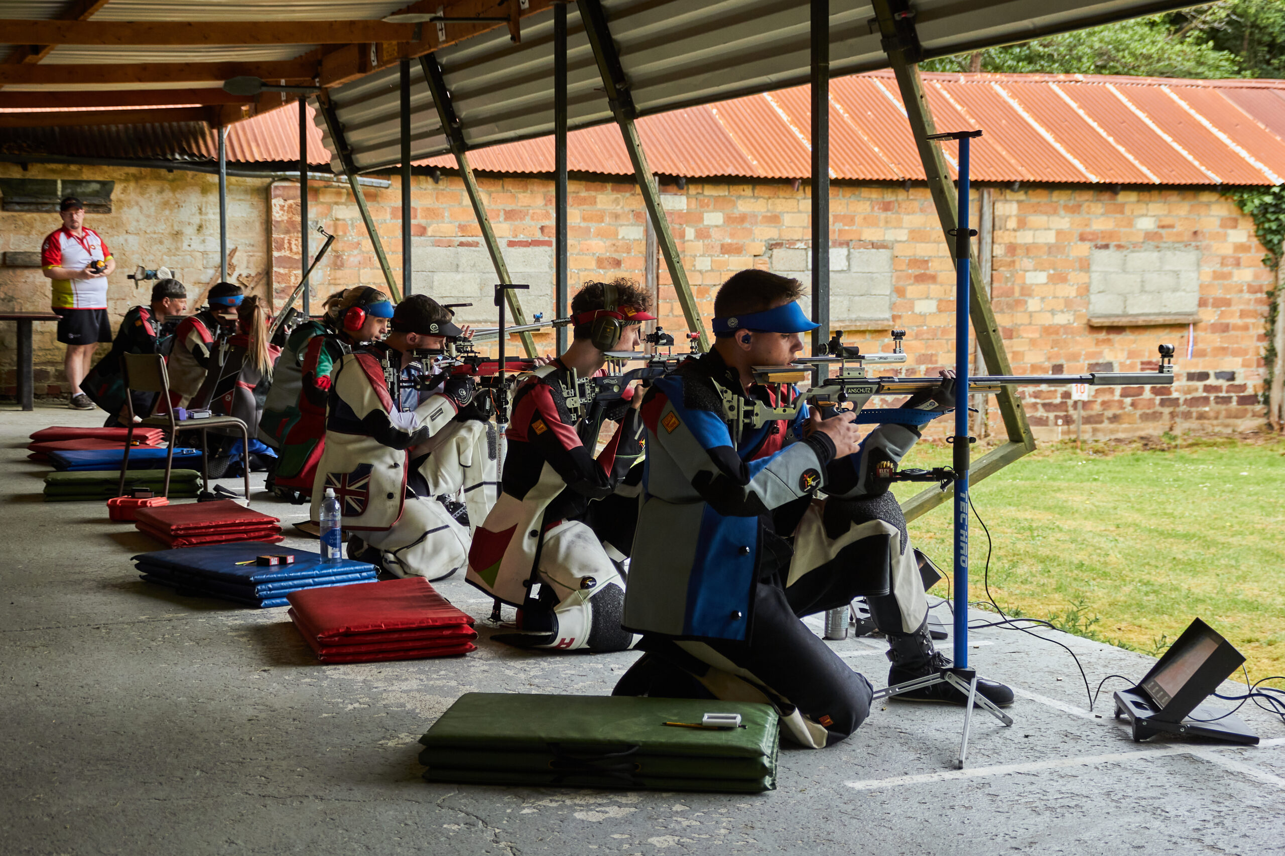 Competitors fire target rifles from a kneeling position. The range is outdoors but the firing point is covered. They wear stiff, multi-coloured jackets and trousers to help support the rifles. Credit: Richard Hemingway