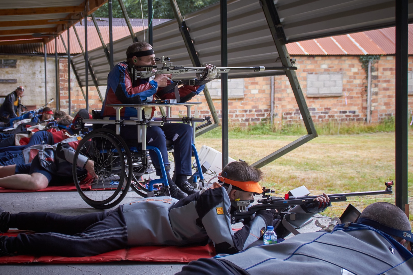 Target shooting competitors take aim on the covered firing point of an outdoor range. In the foreground, a shooter fires lying from the floor. They wear a grey jacket and orange visor. Beyond them, a disabled athlete shoots from a wheelchair. Credit: Richard Hemingway