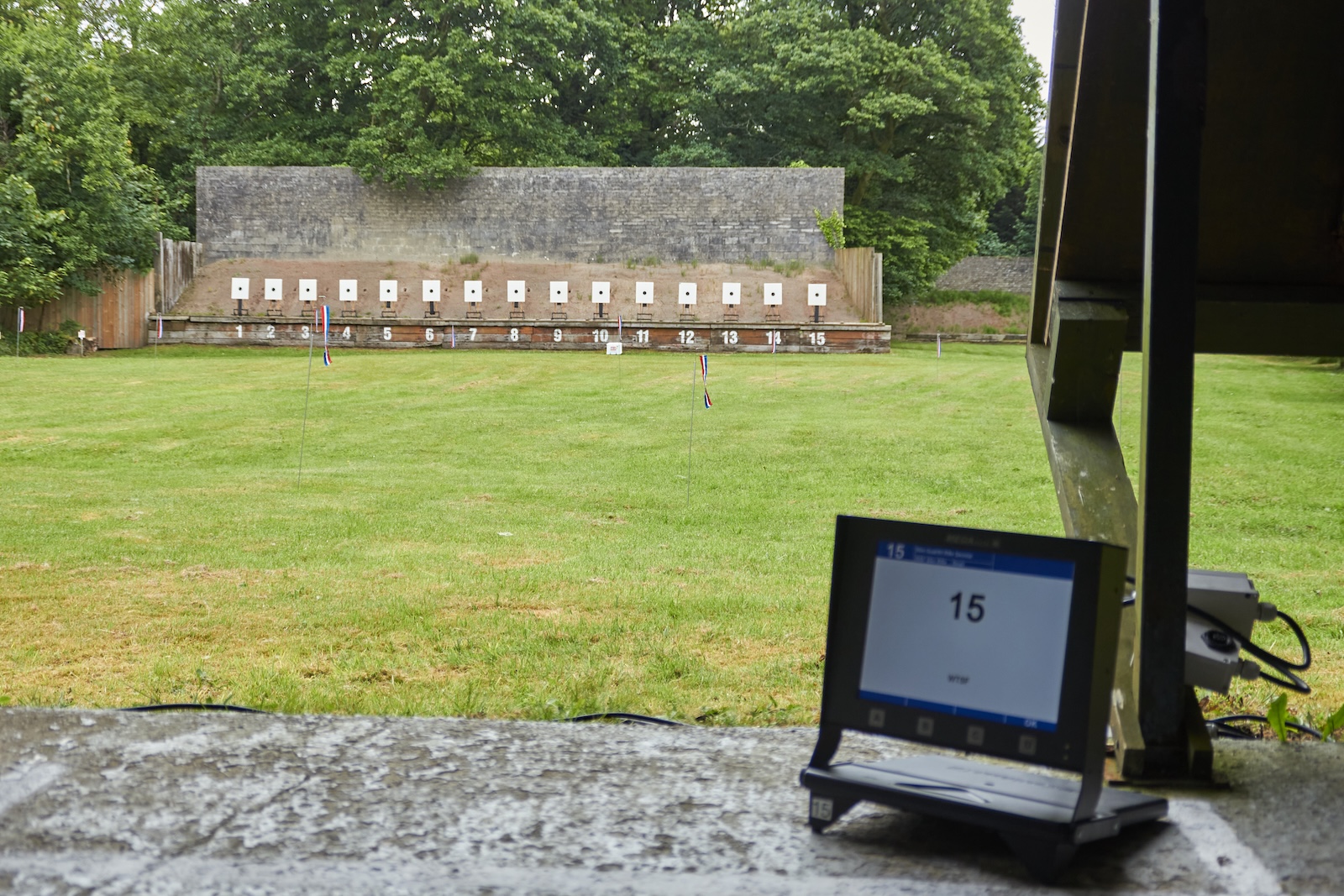 A monitor for an electronic shooting target sits on the firing point of a grassy range. In the distance a bank of targets are visible in front of an earth berm and concrete walll. Credit: Richard Hemingway