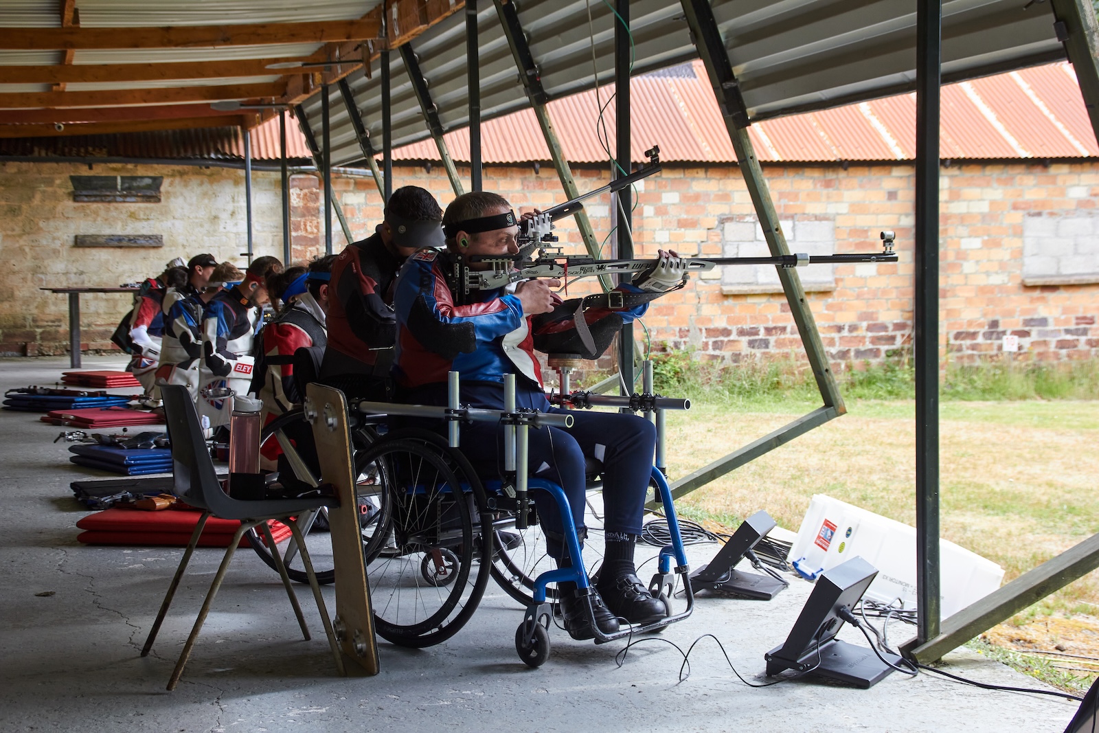 Athletes shoot from a covered firing point on an outdoor shooting range. Nearest the camera are two para athletes shooting from wheelchairs. Credit: Richard Hemingway