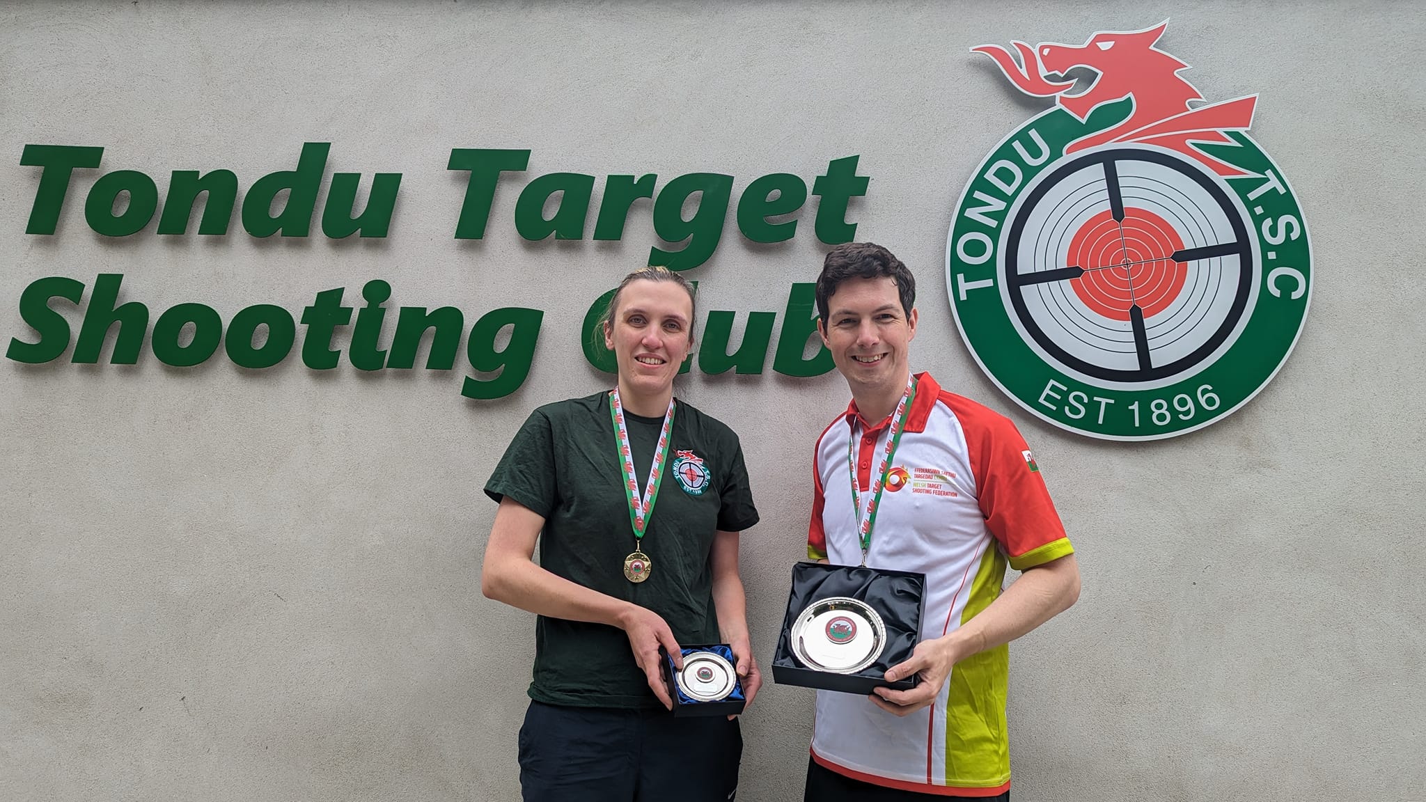 Two people stand in front of a wall bearing a sign that reads "Tondu Target Shooting Club". They are wearing medals and holding silver plates. Credit: Dave Phelps