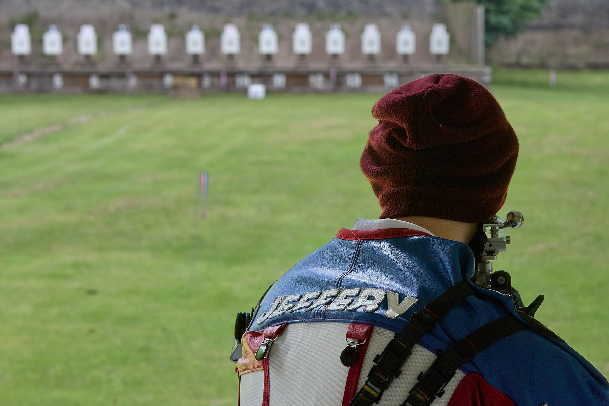A target shooter aims on a shooting range. They wear a red hat and a special blue and white jacket shooting which has the name "Jeffery" stitched onto the back of it.