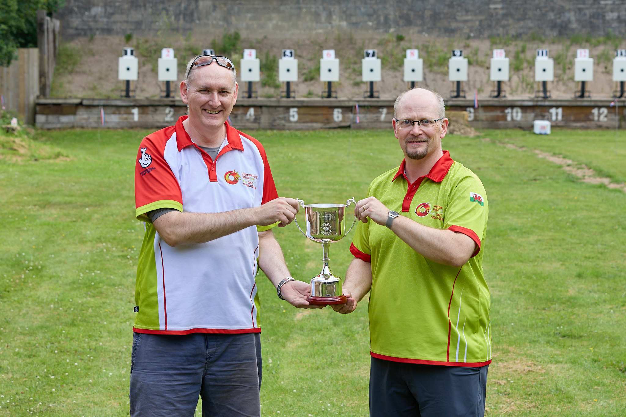 Two people stand on a shooting range holding a trophy