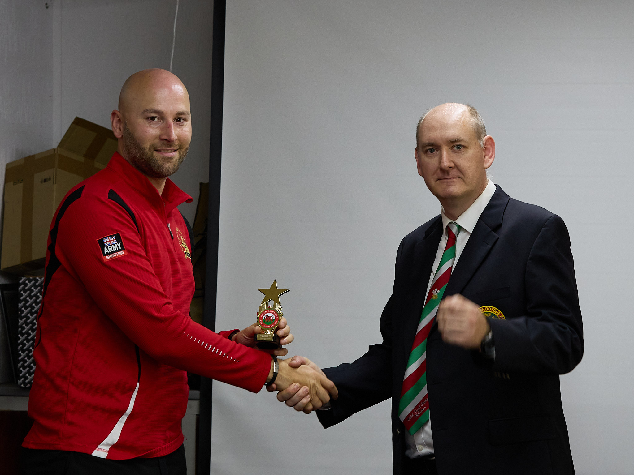 Two people stand in front of a projection screen. The man wearing a formal jacket and tie presents prize to an athlete.