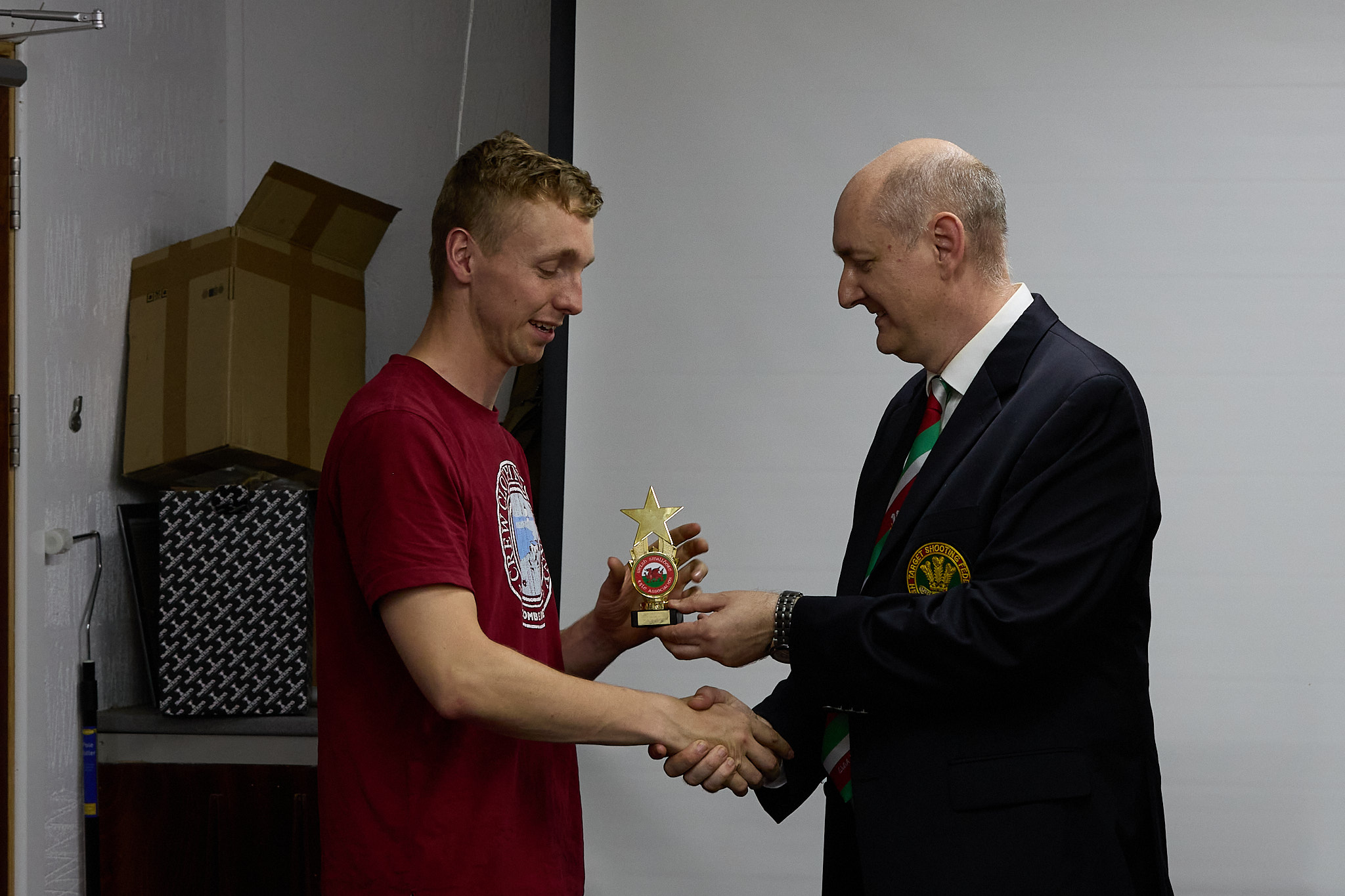 Two people stand in front of a projection screen. The man wearing a formal jacket and tie presents prize to an athlete.