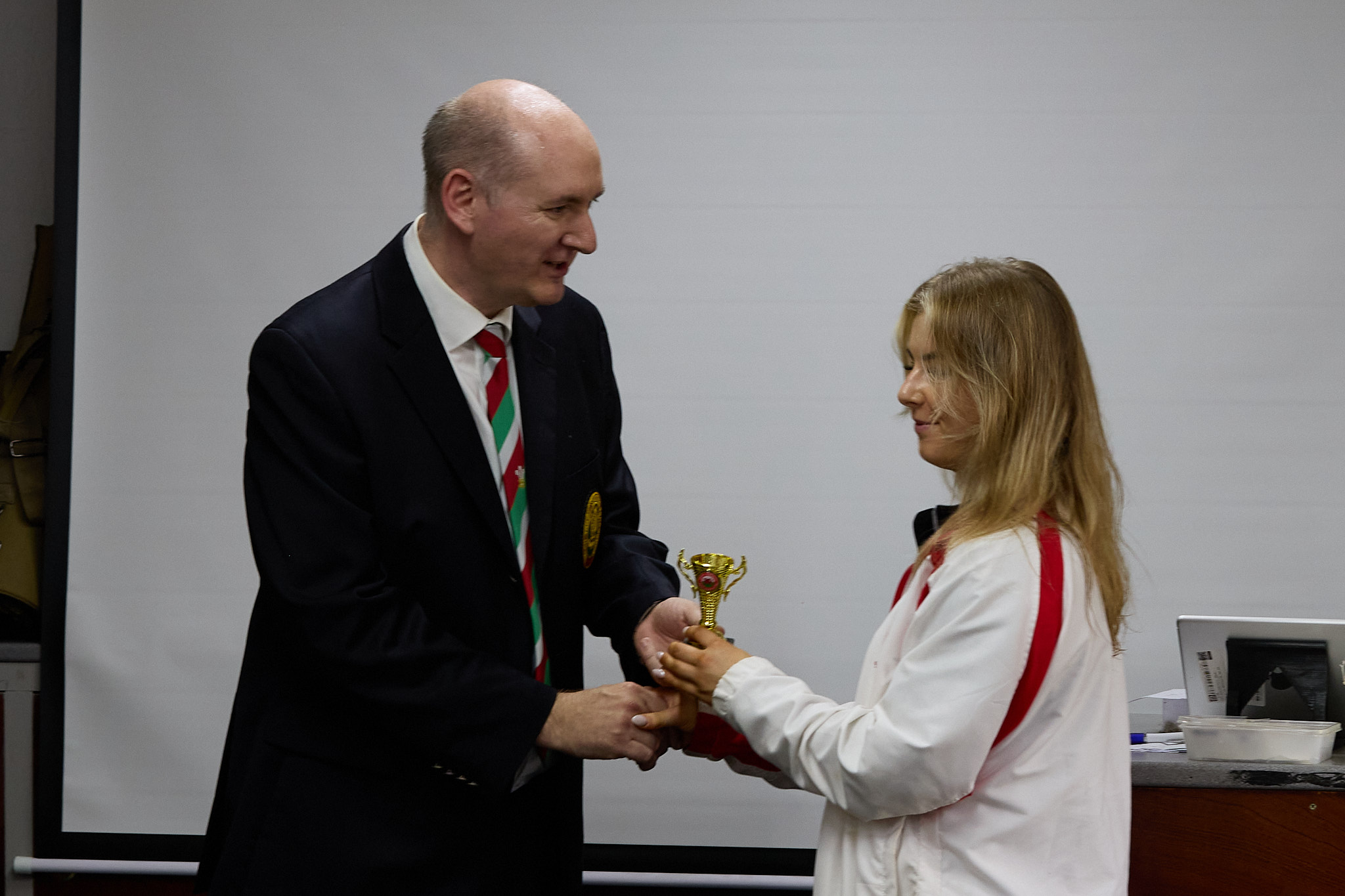 Two people stand in front of a projection screen. The man wearing a formal jacket and tie presents prize to an athlete.