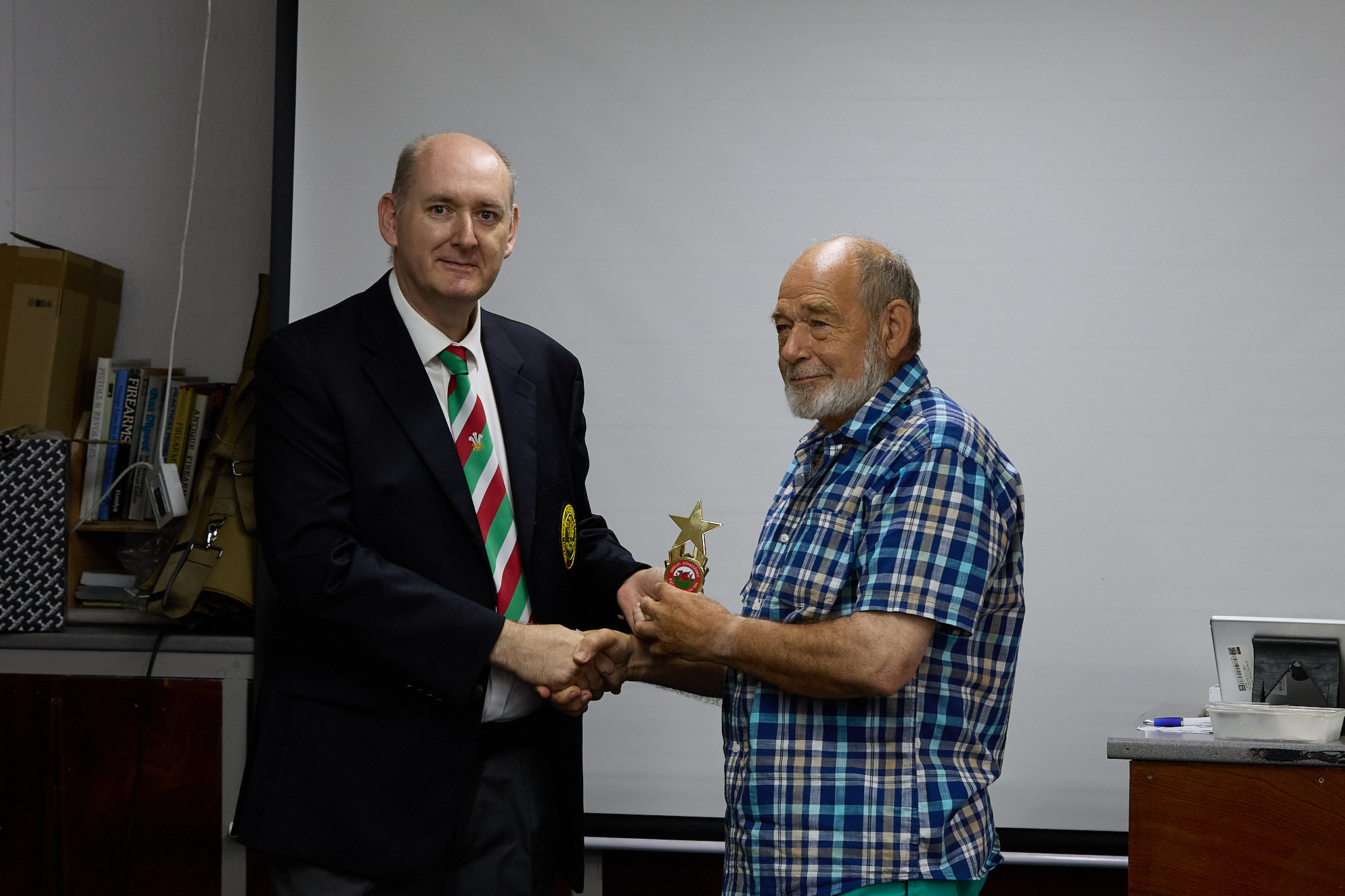 Two people stand in front of a projection screen. The man wearing a formal jacket and tie presents prize to an athlete.