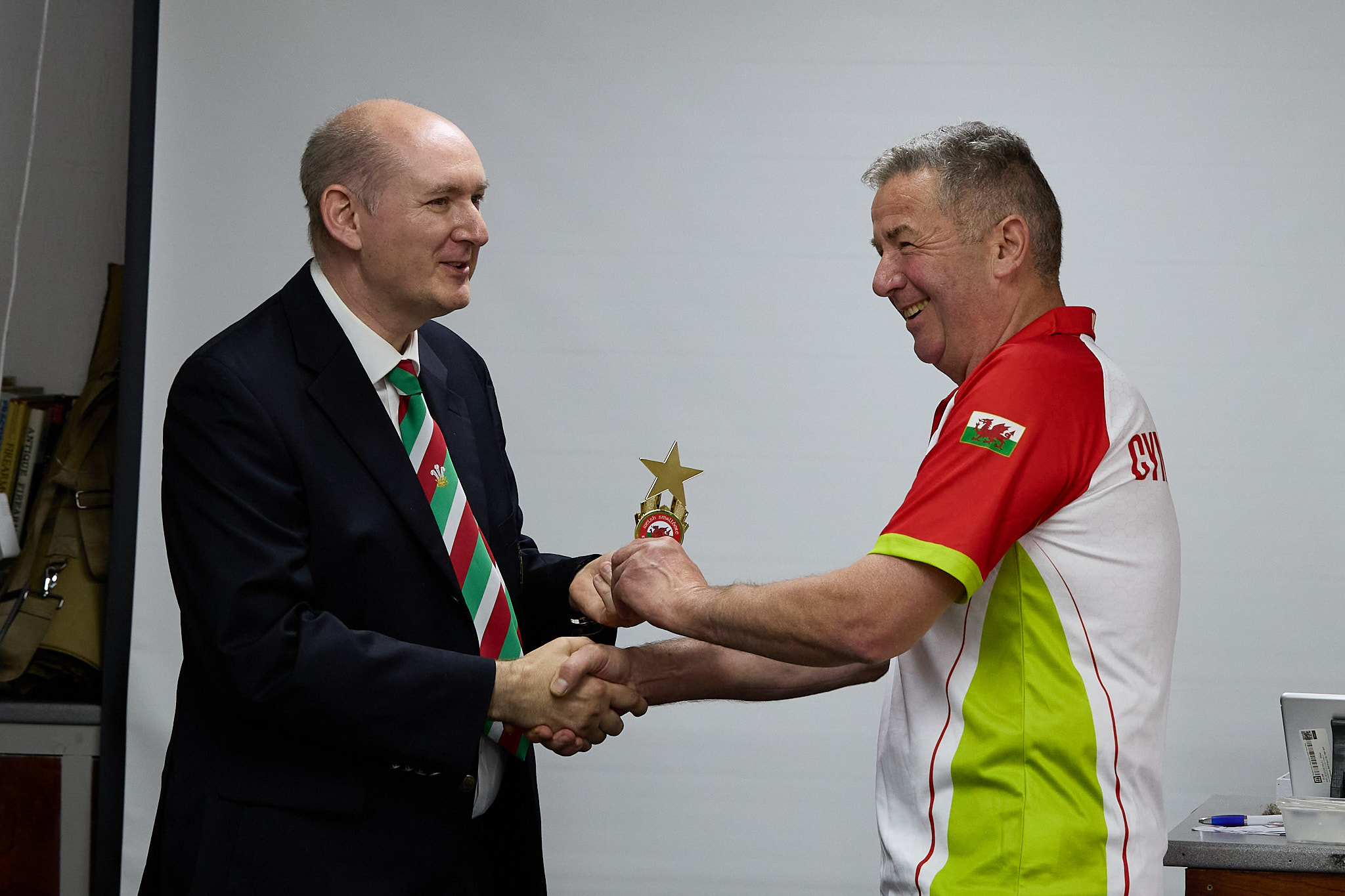Two people stand in front of a projection screen. The man wearing a formal jacket and tie presents prize to an athlete.