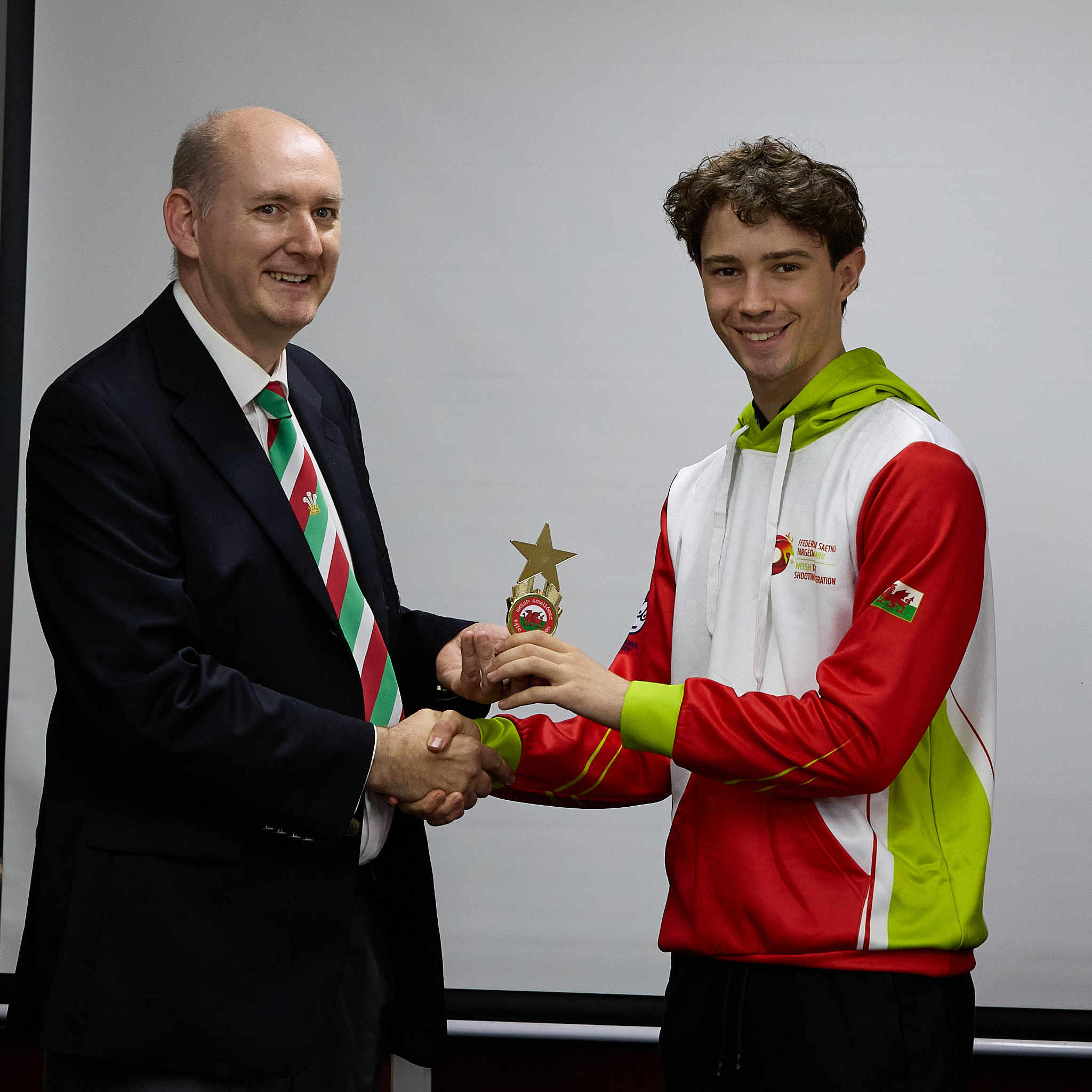 Two people stand in front of a projection screen. The man wearing a formal jacket and tie presents prize to an athlete.