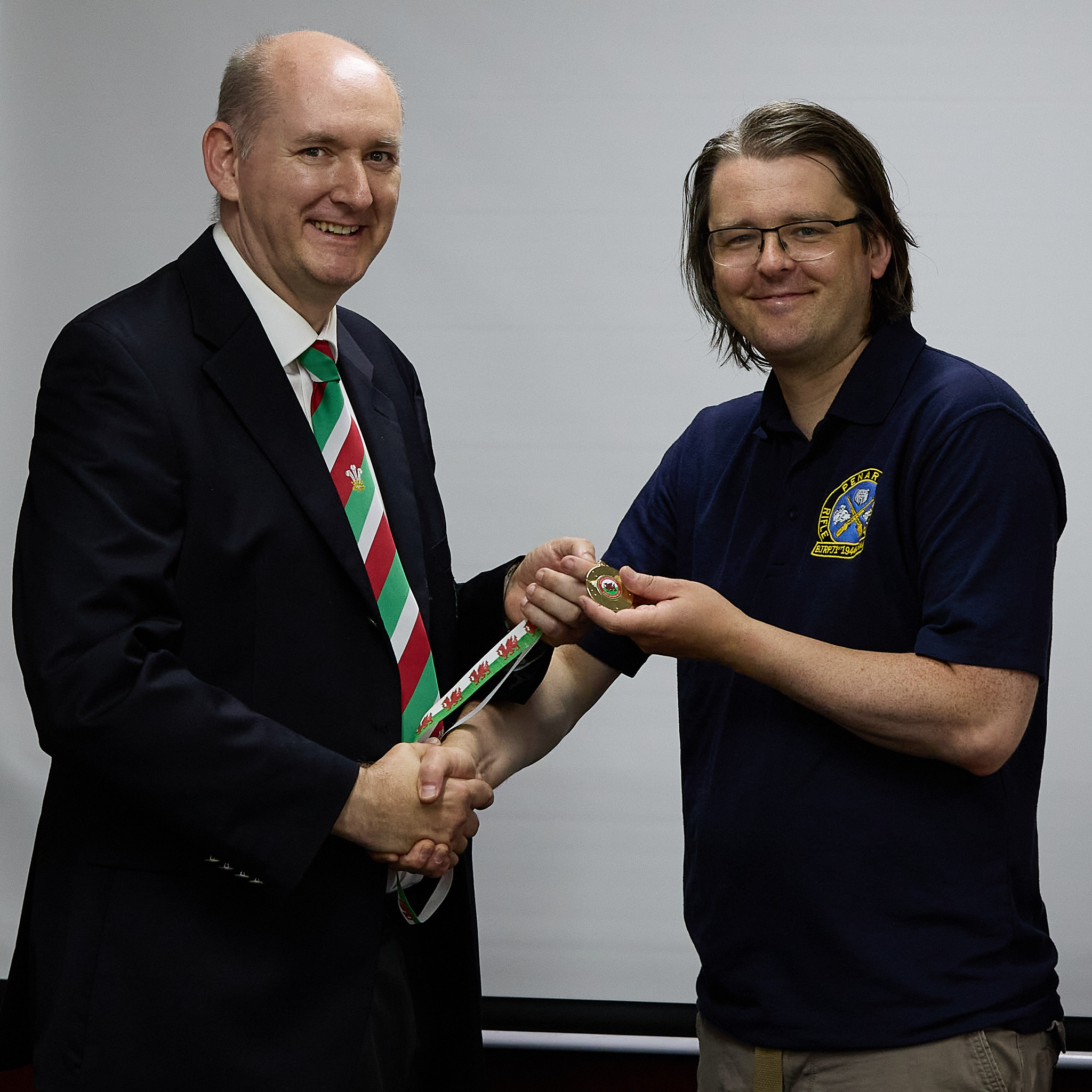 Two people stand in front of a projection screen. The man wearing a formal jacket and tie presents a medal to an athlete.