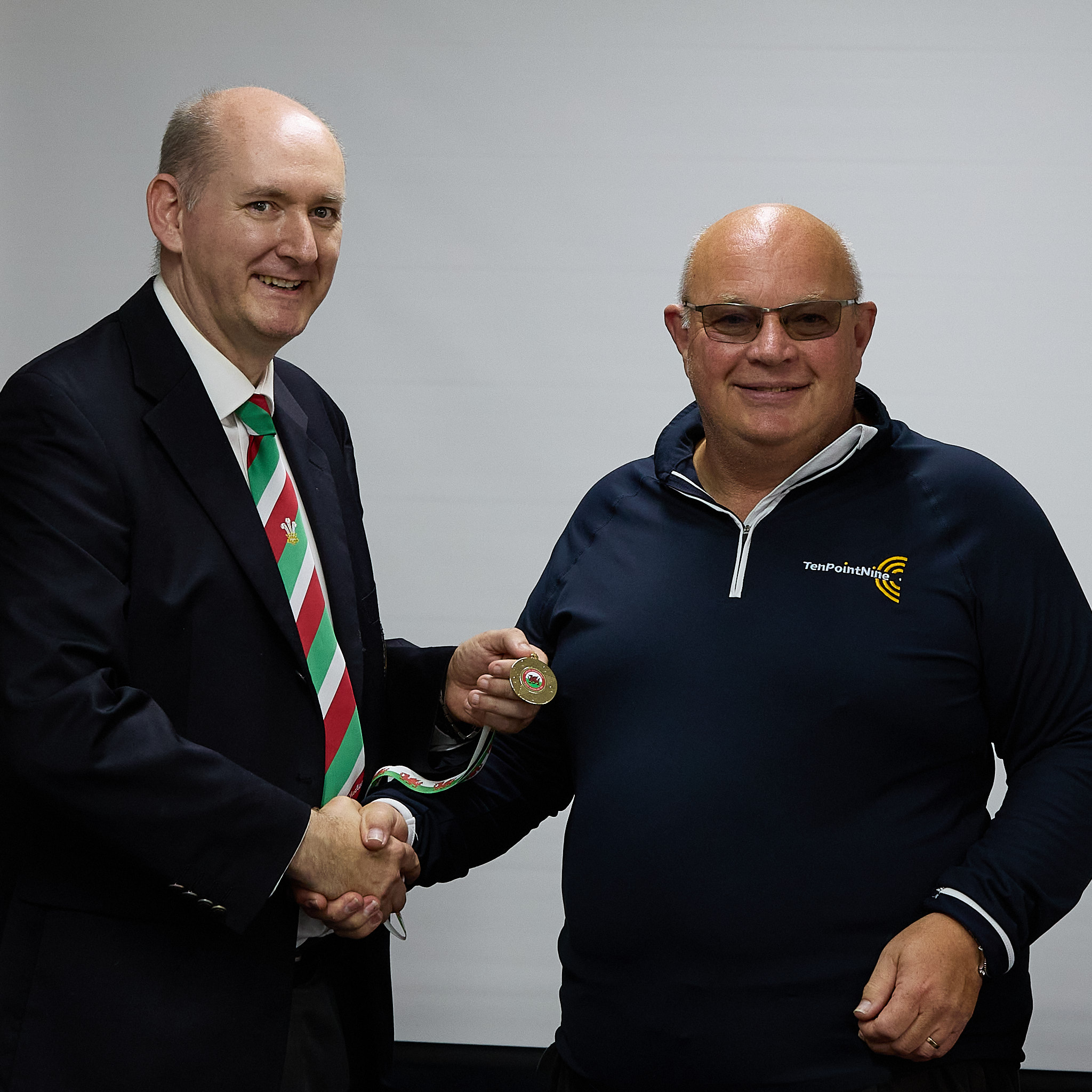 Two people stand in front of a projection screen. The man wearing a formal jacket and tie presents a medal to an athlete.