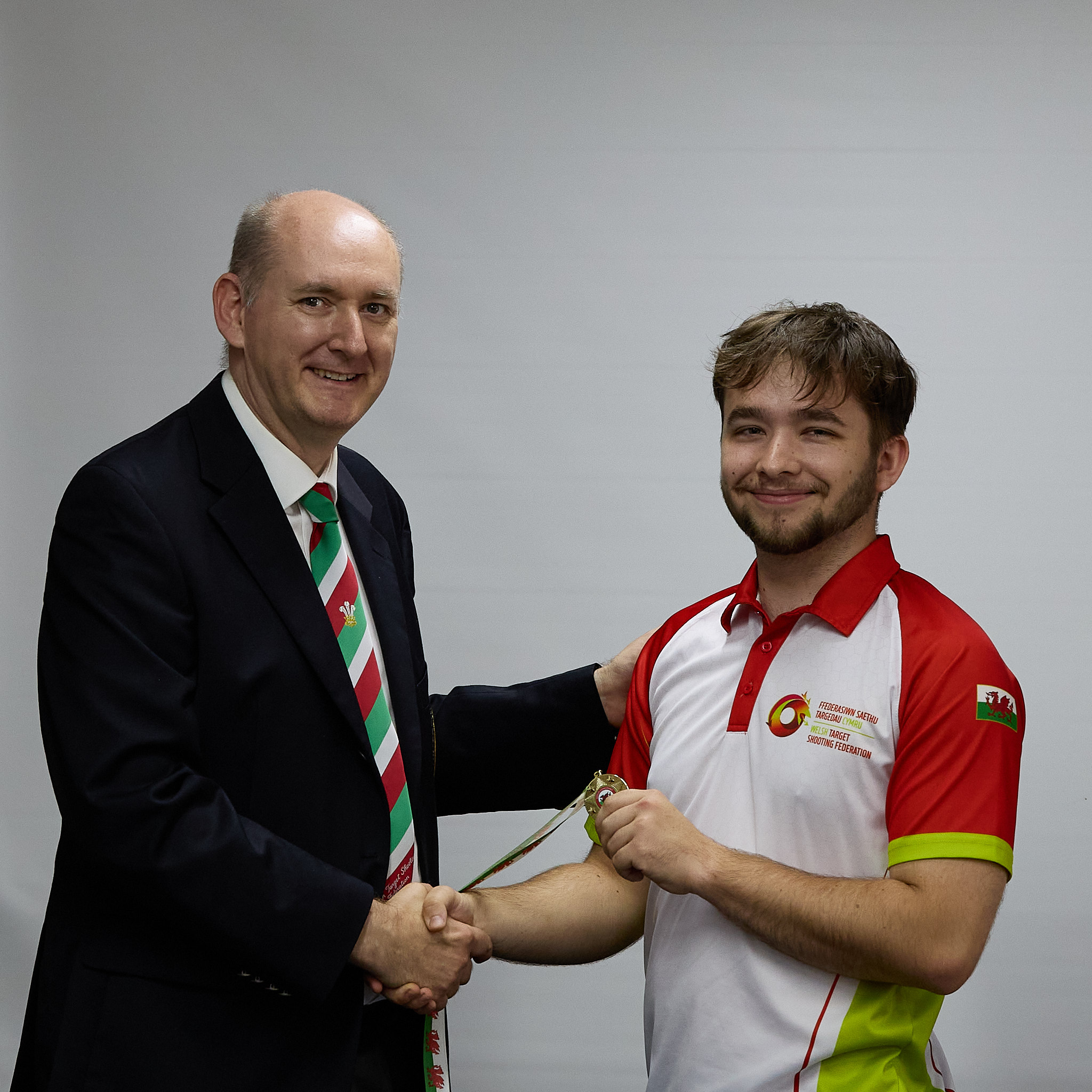 Two people stand in front of a projection screen. The man wearing a formal jacket and tie presents a medal to an athlete.