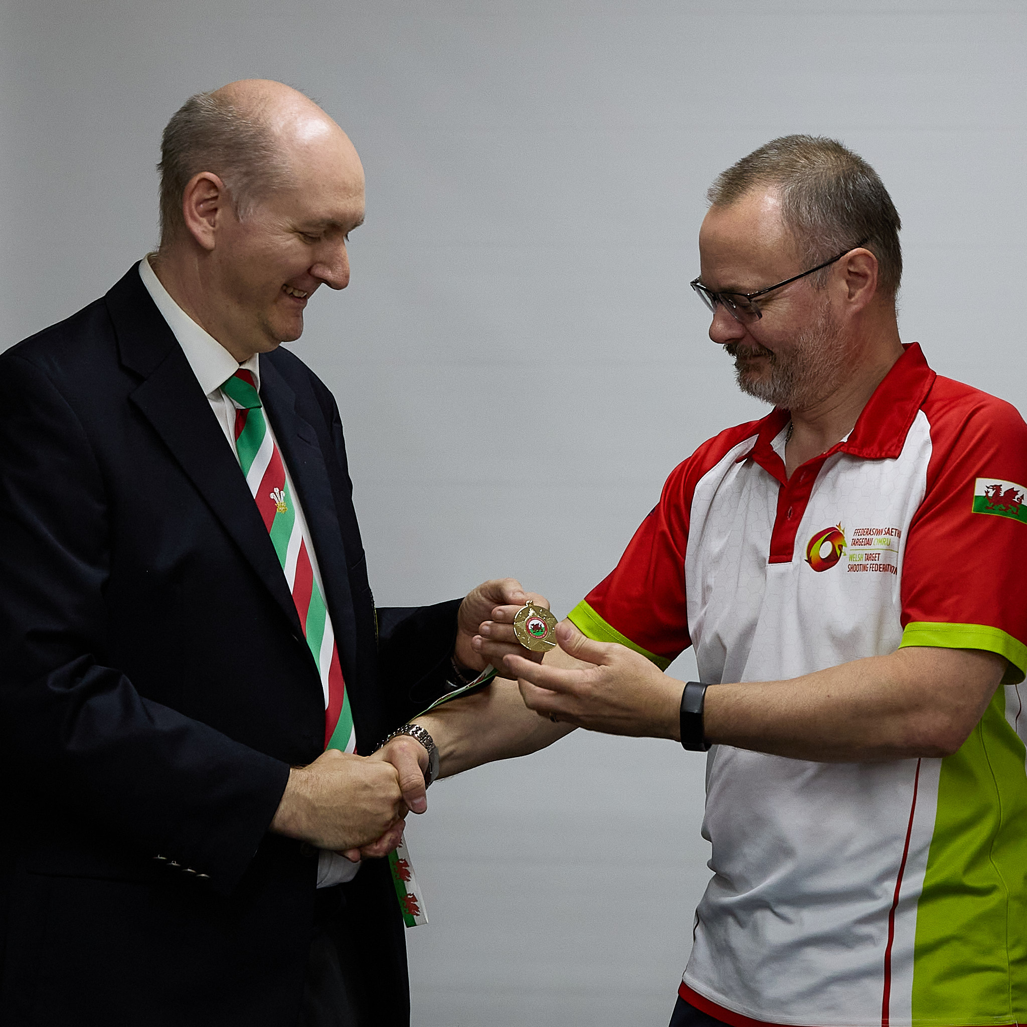 Two people stand in front of a projection screen. The man wearing a formal jacket and tie presents a medal to an athlete.
