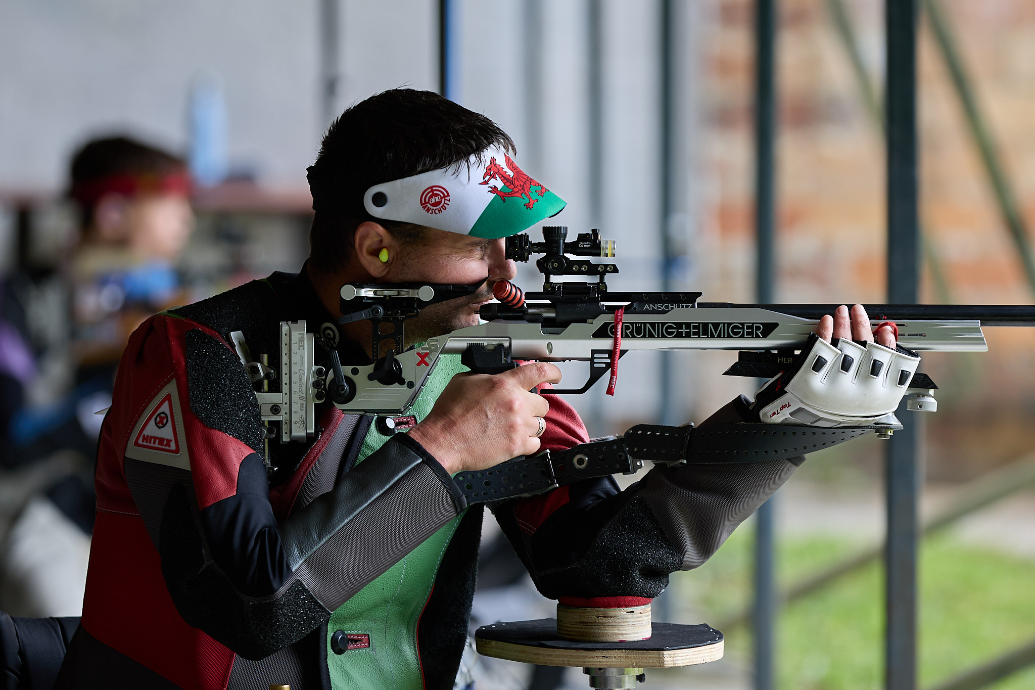 A male Para athlete aims a target rifle from the "kneeling" position. As he is in a wheelchair, this means he allowed to rest his left elbow but the right elbow must be unsupported in free space. He wears visor bearing the Welsh dragon and a red and green shooting jacket.