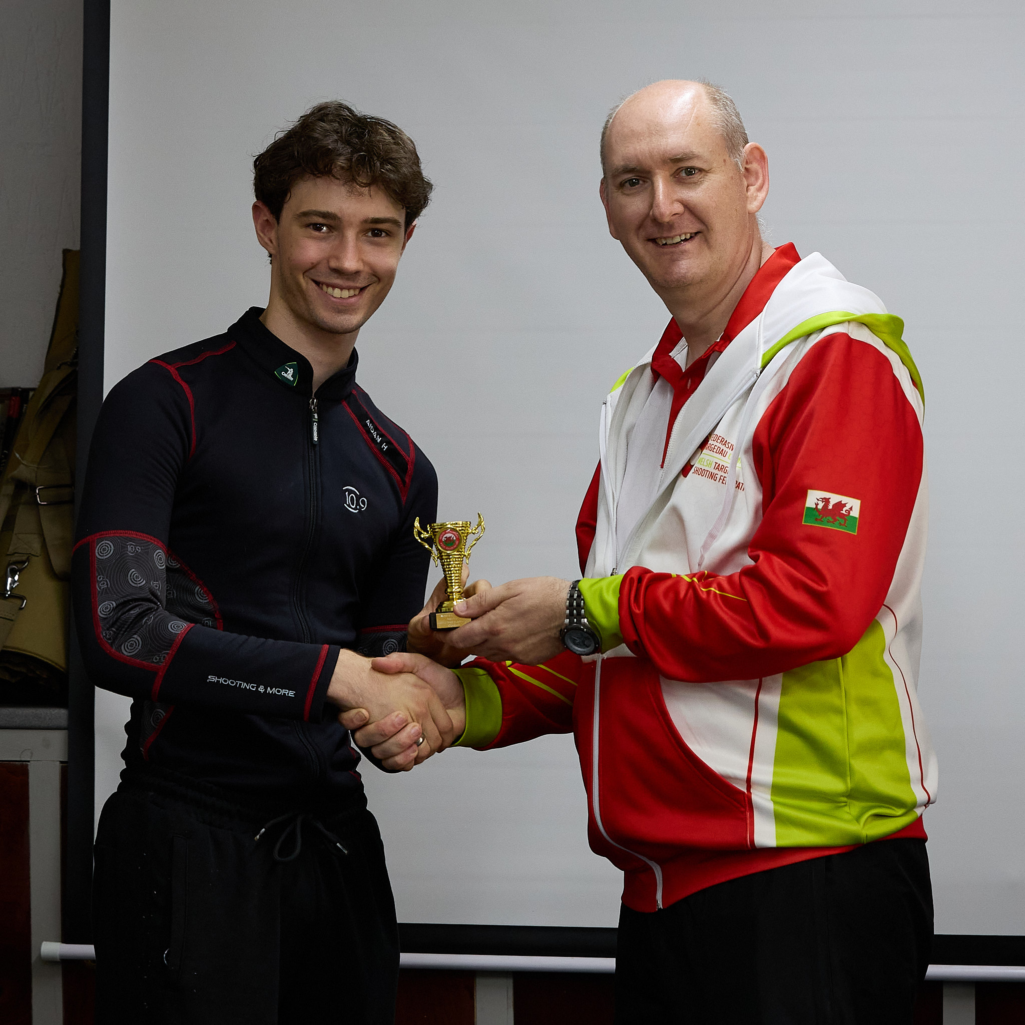 Two people stand in front of a projection screen. The man in a red, white and green top presents a prize to an athlete.