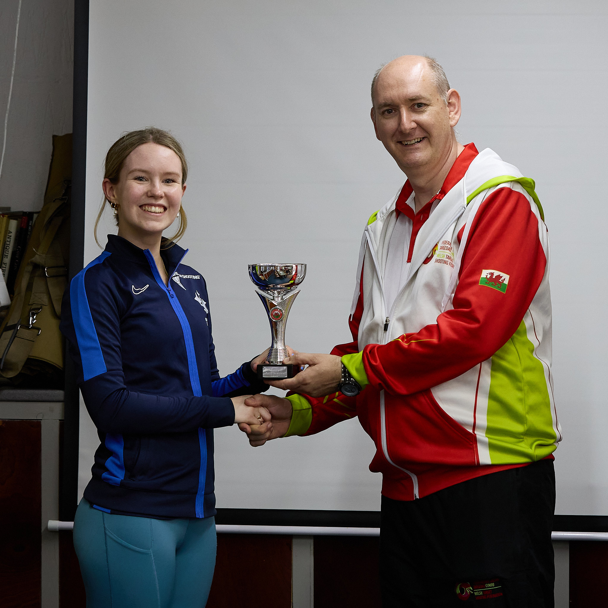 Two people stand in front of a projection screen. The man in a red, white and green top presents a prize to an athlete.