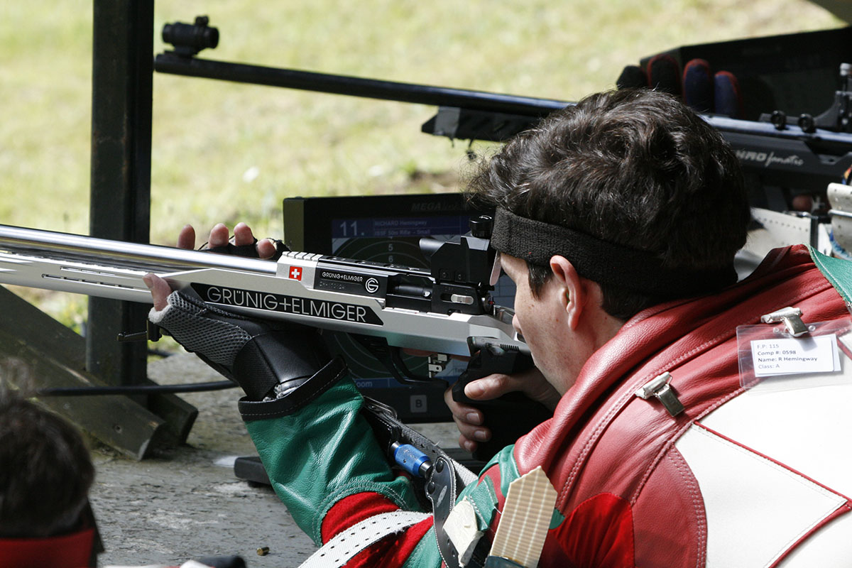 A male athlete takes aim in the prone position with a silver target rifle. He wears a red and green shooting jacket and ablack glove. Credit: Niall Evans