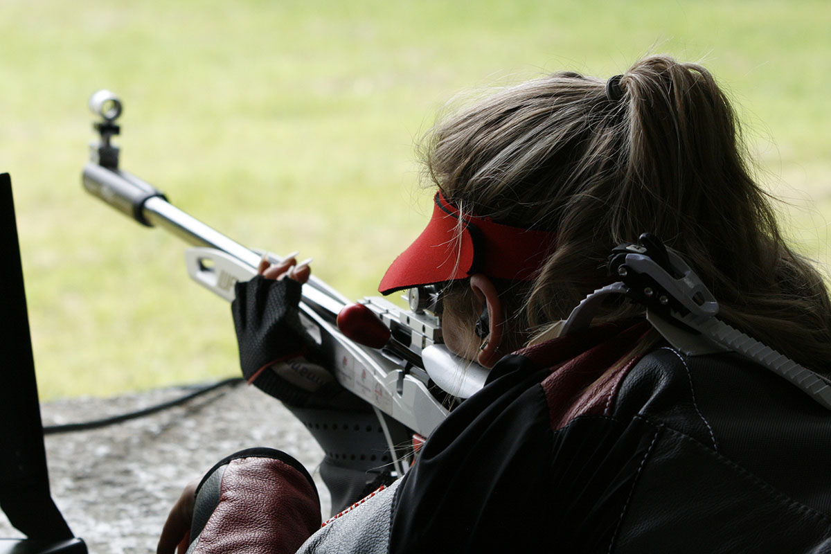 A female athlete takes aim in the prone position with a silver target rifle. She wears a black and grey shooting jacket and a red visor. Credit: Niall Evans