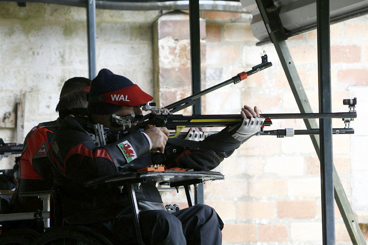 A wheelchair athlete takes aim with a black target rifle. They wear a red and black shooting jacketand a red visor. A welsh flag is visible on their sleeve. Credit: Niall Evans