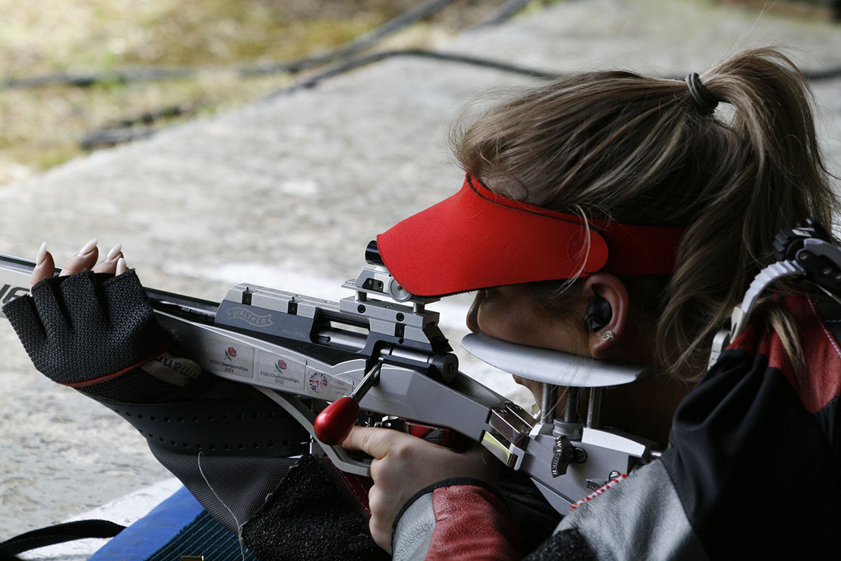 A female athlete takes aim in the prone position with a silver target rifle. She wears a black and grey shooting jacket and a red visor. Credit: Niall Evans