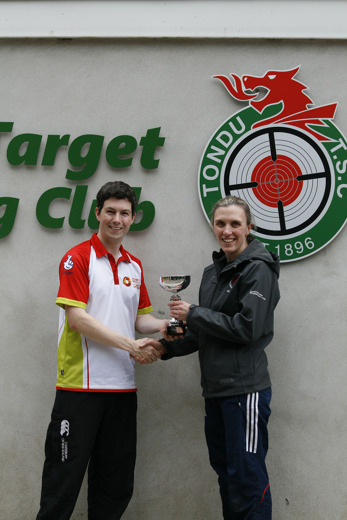 A man in a red and white top presents a trophy to a female athlete in a grey jackets and black tracksuit bottoms. Credit: Niall Evans