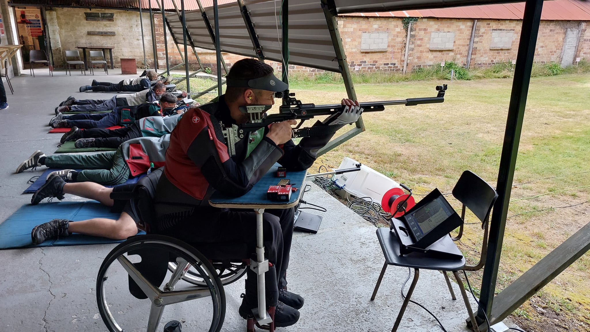 A target shooter in a wheelchair takes aim with a black target rifle on an outdoor range. Behind them other competitors shoot from the floor in the prone position. Credit: Steve Gurner