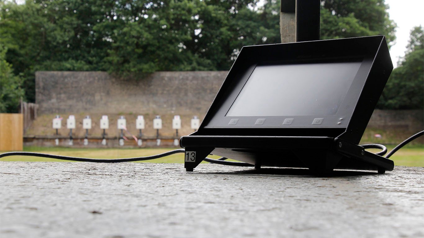 A target monitor sits on the firing point of an outdoor shooting range. A row of rifle targets is visible in the distance in front of a sandbank and protective wall. Credit: Niall Evans