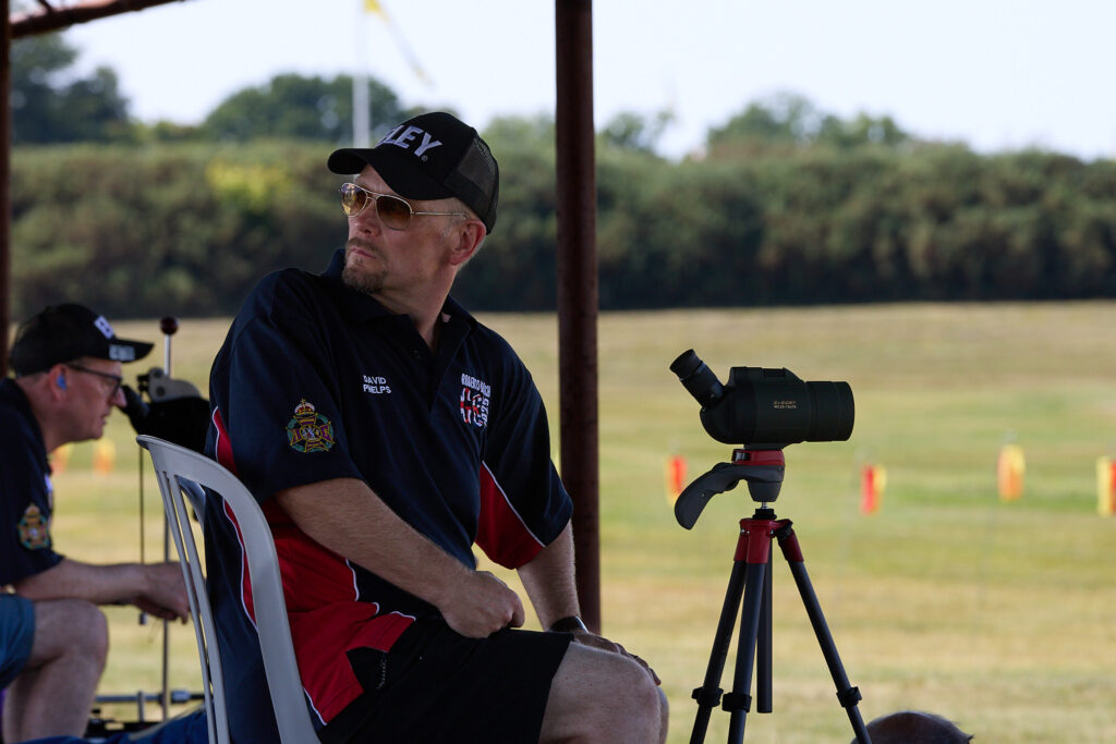 A man sits on a plastic chair on a shooting range firing point. He looks over this right shoulder to see the behaviour of wind flags behind him and any incoming gusts. A scope is mounted in front of him on a tripod, allowing him to watch the movement of mirage. Blurred red and yellow wind flags are visible in the distance.