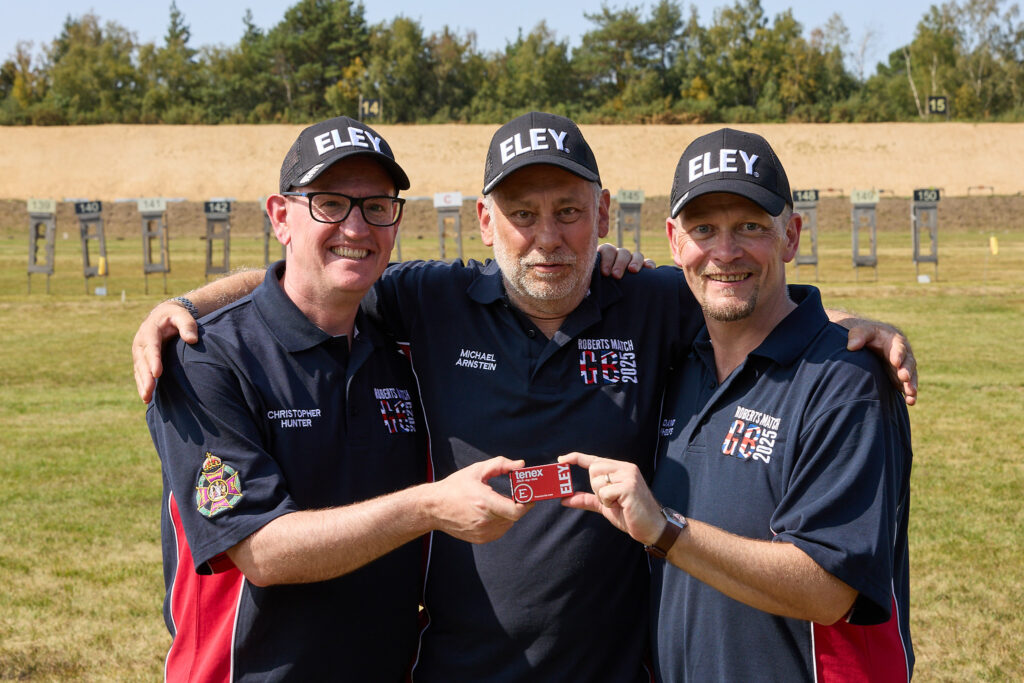 Three smiling men stand on a shooting range. They wear black caps with the word "Eley" on it, and hold a box of Eley Tenex match ammunition. They wear shirts bwith the words "Roberts Match GB 2025" on.