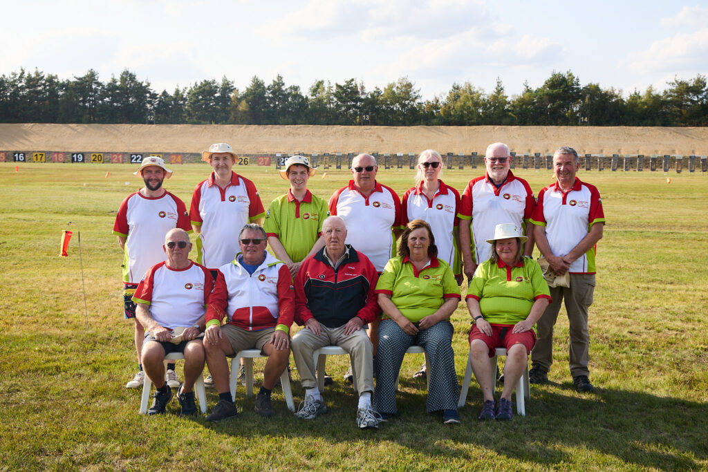 A group of smiling people pose for a group photograph on a shooting range. The three at the front are seated. They wear tops bearing the emblem of the Welsh Target Shooting Federation.
