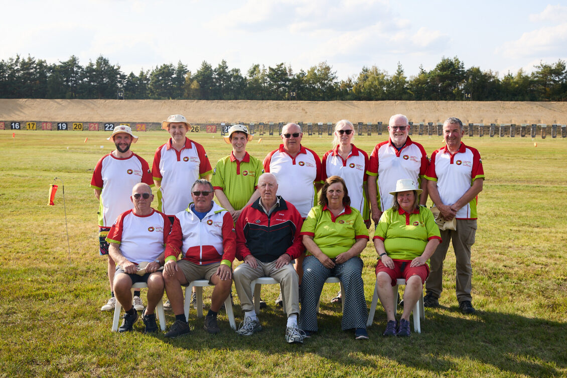 A group of smiling people pose for a group photograph on a shooting range. The three at the front are seated. They wear tops bearing the emblem of the Welsh Target Shooting Federation.