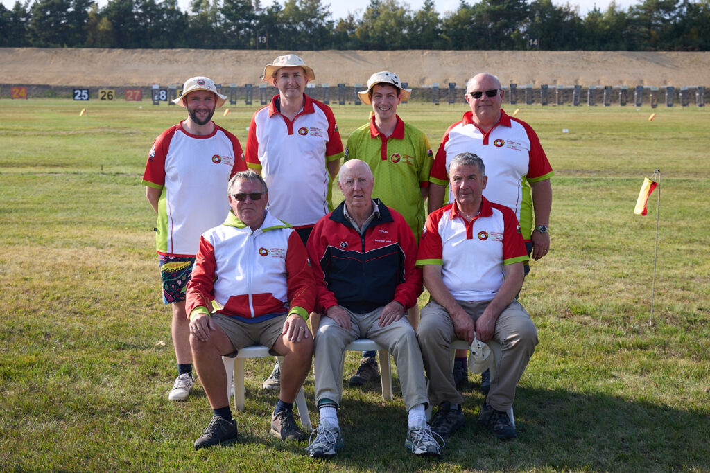 A group of smiling men pose for a group photograph on a shooting range. The three at the front are seated. They wear tops bearing the emblem of the Welsh Target Shooting Federation.