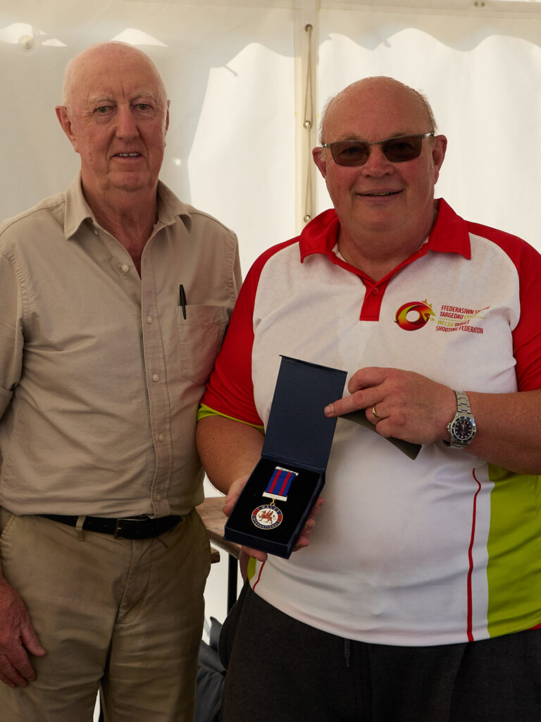 Two smiling men stand in a tent, one is holding a medal they have been awarded. It bears a red welsh dragon and the words "Welsh Shooting".