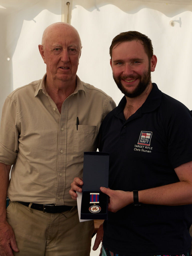 Two smiling men stand in a tent, one is holding a medal they have been awarded. It bears a red welsh dragon and the words "Welsh Shooting".