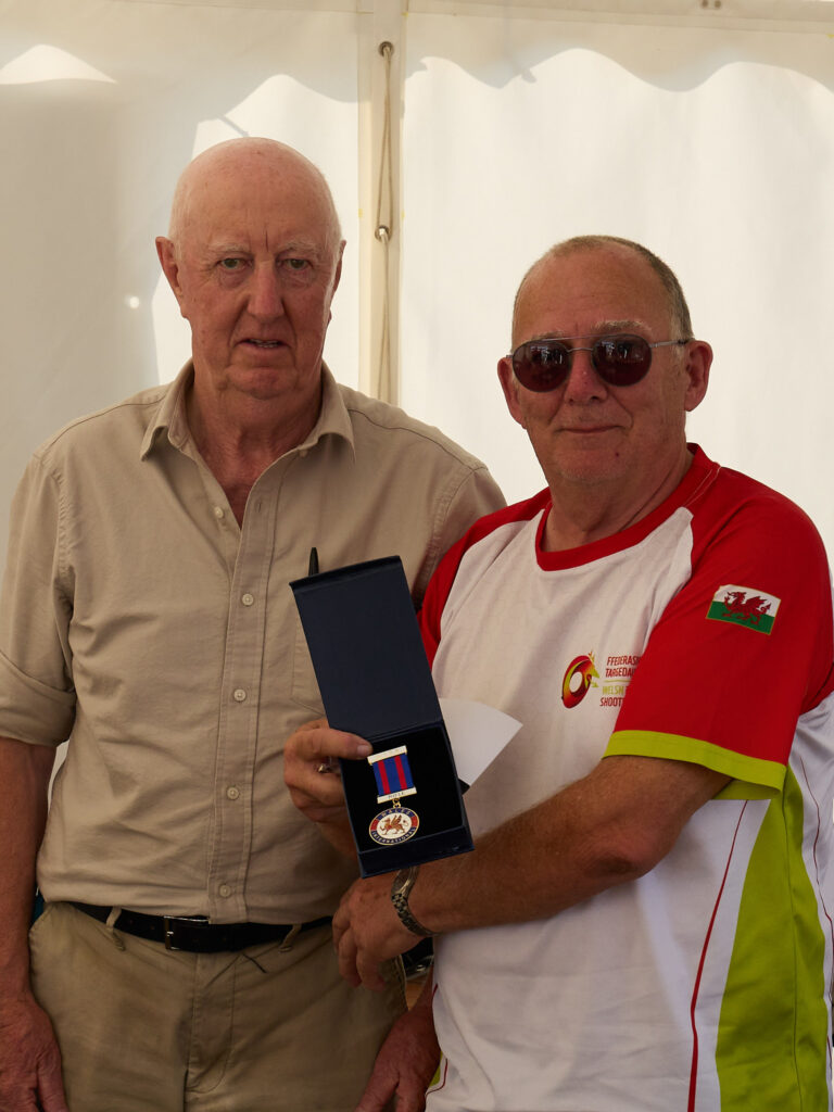Two smiling men stand in a tent, one is holding a medal they have been awarded. It bears a red welsh dragon and the words "Welsh Shooting".