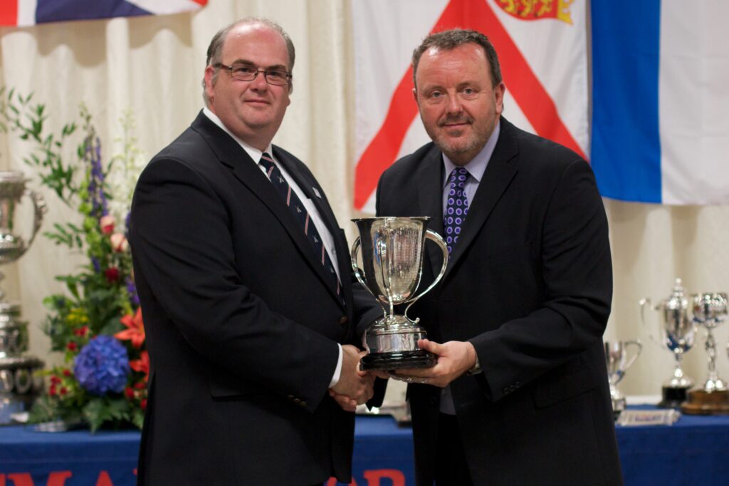 Two smiling men pose in front of a table laden with silverware as the one on the right presents a trophy to the man on the left.