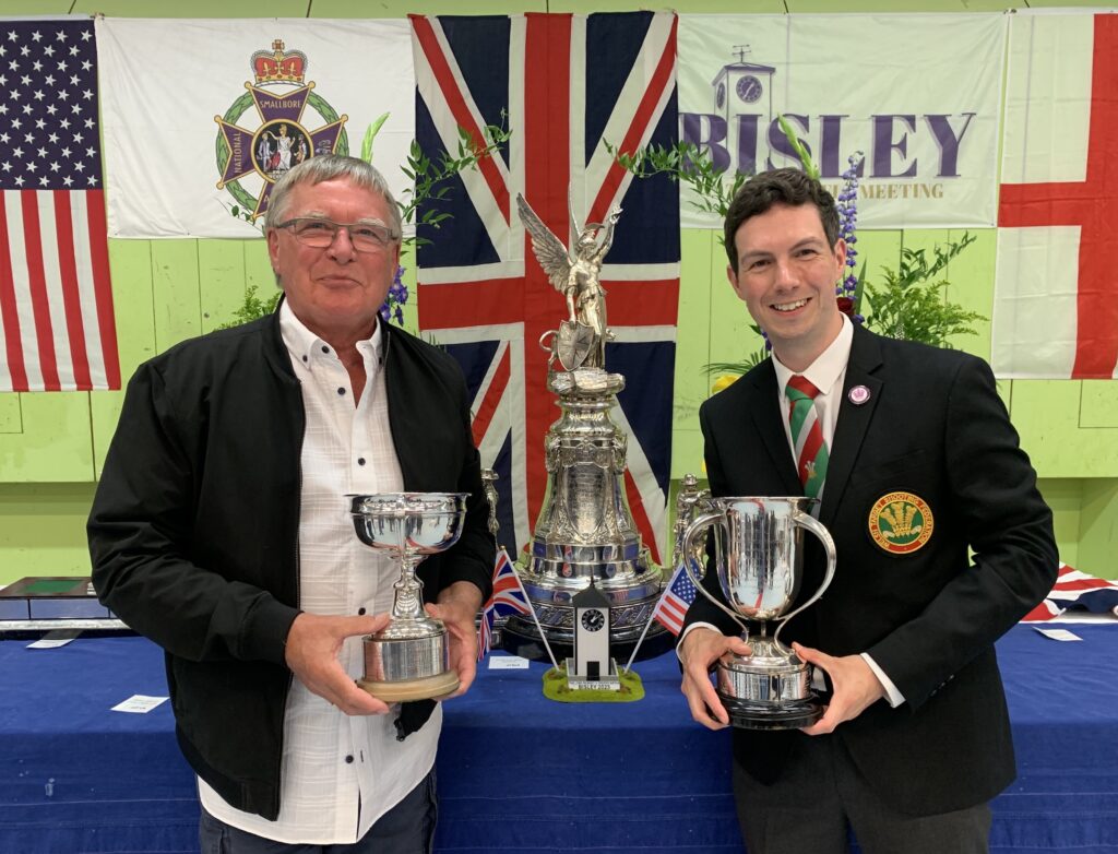 Two smiling men stand holding silver trophies. One is in a casual white shirt and black jacket. The other is suited with a red and green tie and a black blazer bearing a red and green badge. The badge reads "Welsh Target Shooting Federation" and is embroidered with the Prince of Wales Feathers. They stand in front of a table covered in a blue cloth, on which stands a huge silver trophy. Behind the table the Union flag and other flags are hung.