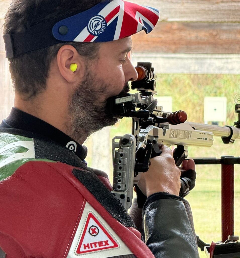 A man holds a rifle in his hands. He wears a visor bearing a Union Flag motif and a red leather shooting jacket.