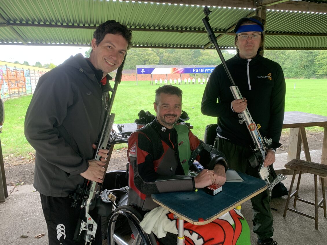 Three smiling men pose on the firing point of a rifle range. The one in the middle sits in a wheelchair with a special table attached. He wears a red and green shooting jacket and has a blanket over his legs bearing the motif of the Welsh flag. The two either side hold smallbore target rifles and are dressed in black and grey.