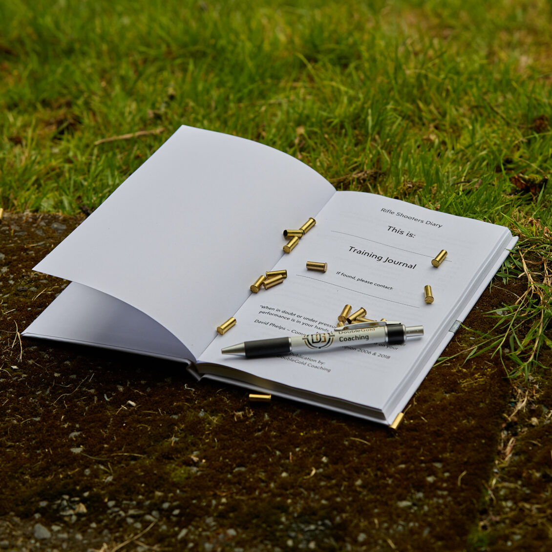 A hardback book lies open on the ground outdoors. A pen and empty cartridge cases lie on the open page, which is titled "Training Journal".