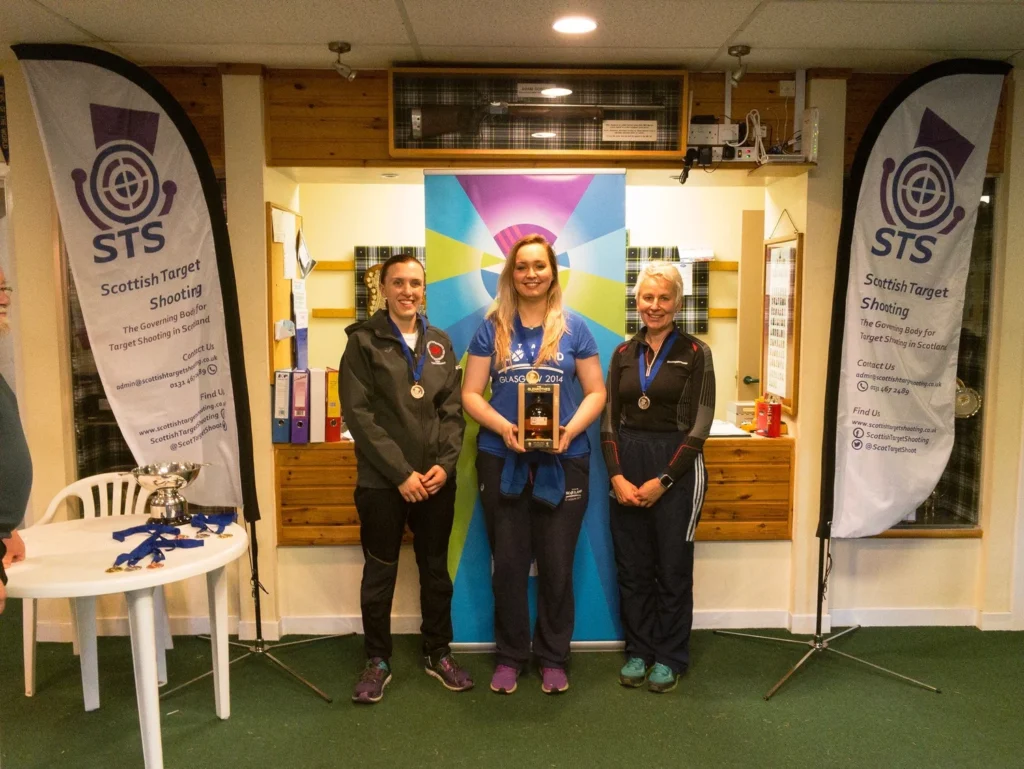 Three women stand in a room with medals. The one in the centre holds a box of whisky. Banners either side read "Scottish Target Shooting".