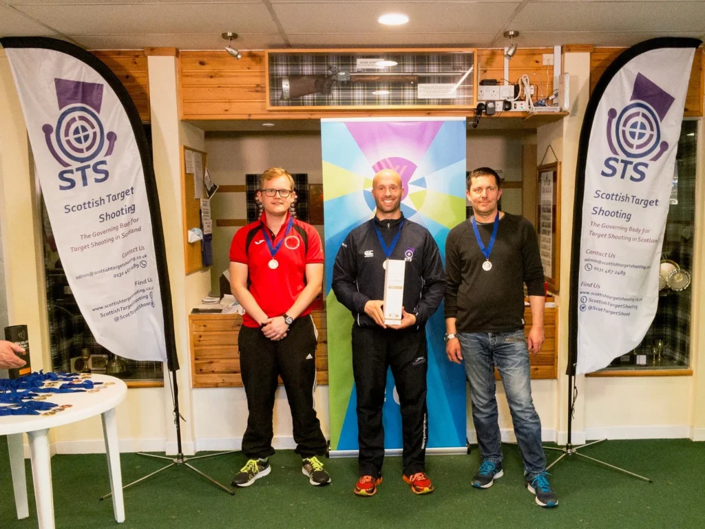 Three men stand in a room with medals. The one in the centre holds a box of whisky. Banners either side read "Scottish Target Shooting".