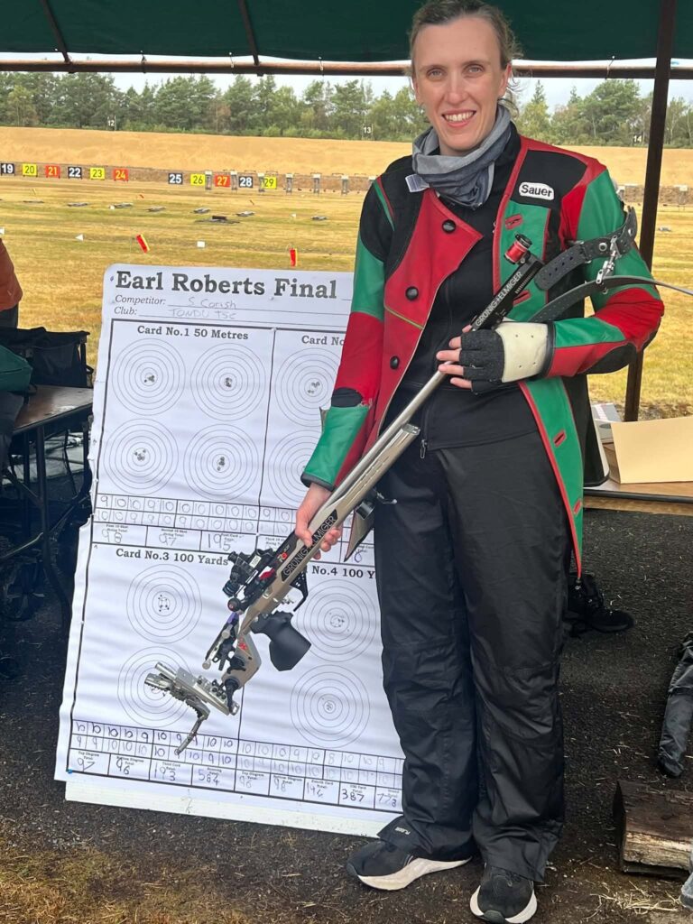A smiling woman stands in front of a score board. She wears a specialist red and green shooting jacket and is holding a silver target rifle bearing the name of Grunig + Elmiger. A grassy outdoor range is visible behind her beyond the covered firing point.
