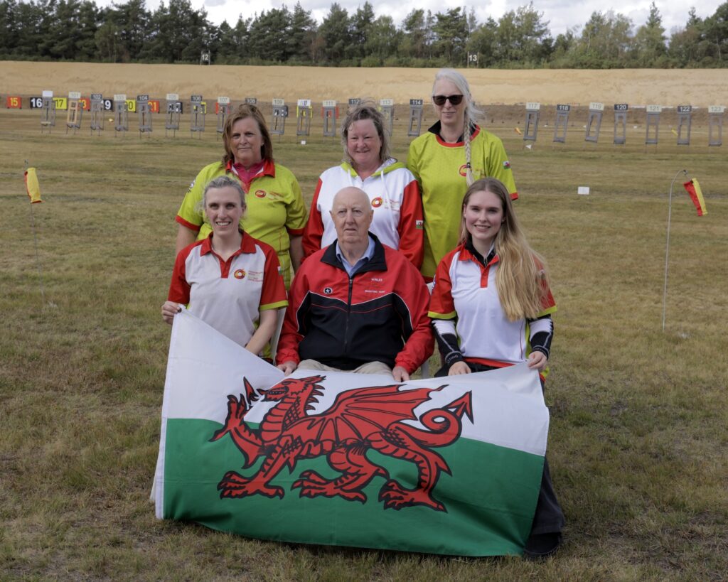 A smiling group of people pose for a team photograph on an outdoor shooting range. They are dressed in red and white team colours and hold a Welsh flag bearing a red dragon on a field of white and green.