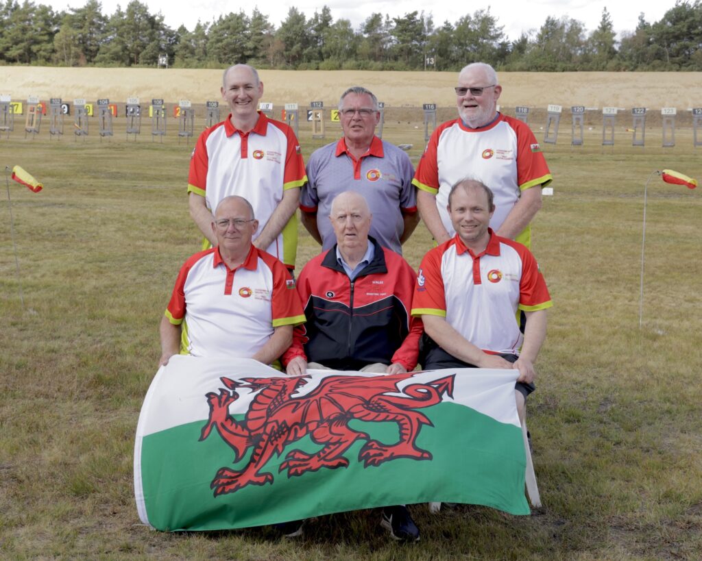 A smiling group of people pose for a team photograph on an outdoor shooting range. They are dressed in red and white team colours and hold a Welsh flag bearing a red dragon on a field of white and green.